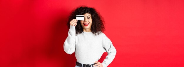 Chica elegante con cabello rizado que muestra una tarjeta de crédito de plástico sobre el ojo y una sonrisa de pie contra la red b