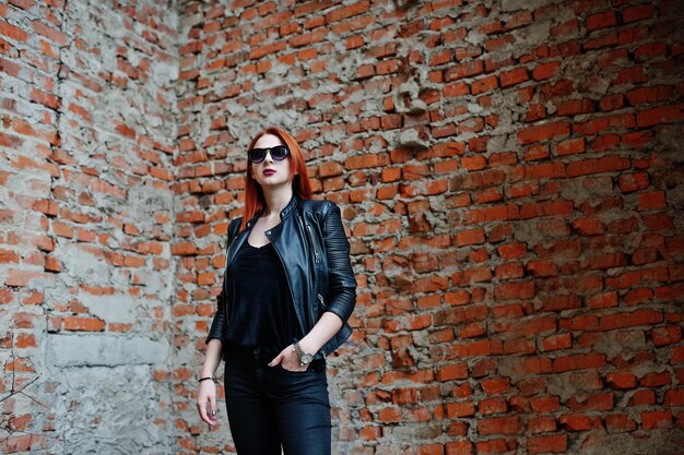 Chica elegante de cabello rojo con gafas de sol vestidas de negro contra un lugar abandonado con paredes de ladrillo