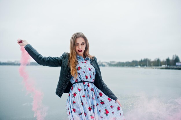 Chica elegante con chaqueta de cuero sostiene una bengala de humo rosa en el día de invierno contra el lago congelado