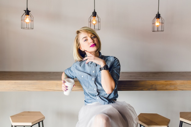 Chica elegante de ensueño con cabello rubio y labios rosados, sentada en una cafetería con mesa y sillas de madera. Ella sostiene una taza de café