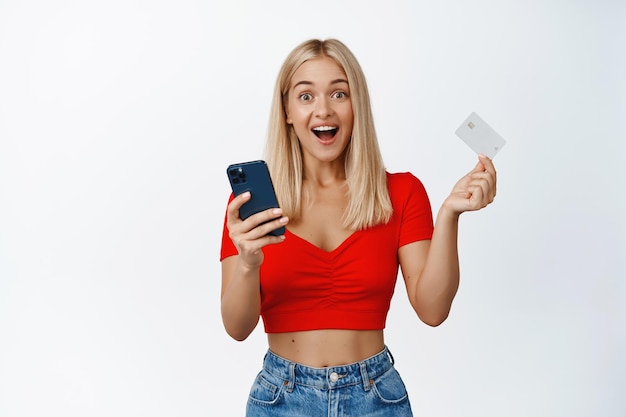 Una chica elegante y feliz hace un pedido en línea con un teléfono y una tarjeta de crédito de fondo blanco