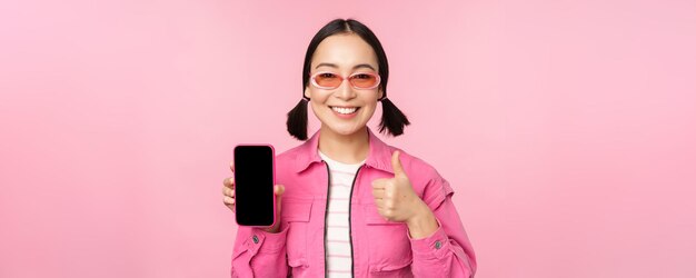 Chica elegante feliz recomienda la aplicación en el teléfono móvil Modelo femenino asiático sonriente que muestra la pantalla del teléfono inteligente y el pulgar hacia arriba de pie sobre fondo rosa