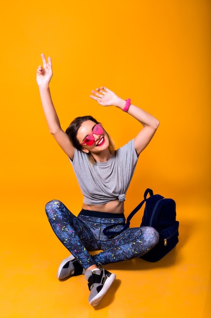 Chica elegante con gafas brillantes con mochila sentada en el suelo con los ojos cerrados. Retrato de estudio de jovencita en calzado deportivo y leggings aislado sobre fondo amarillo.