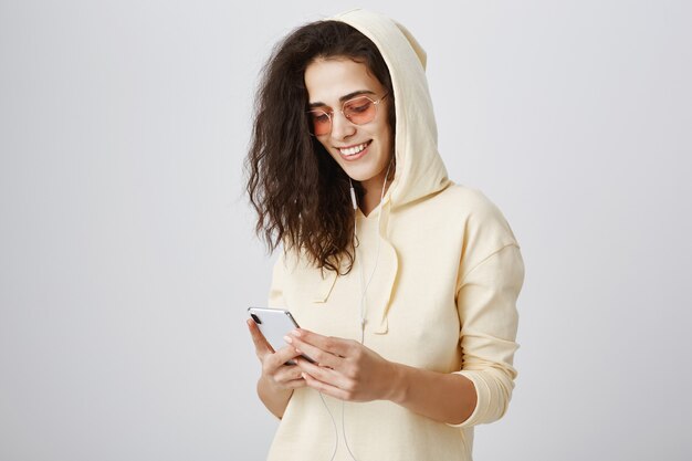 Chica elegante en gafas de sol enviando mensajes de texto a través del teléfono móvil y sonriendo