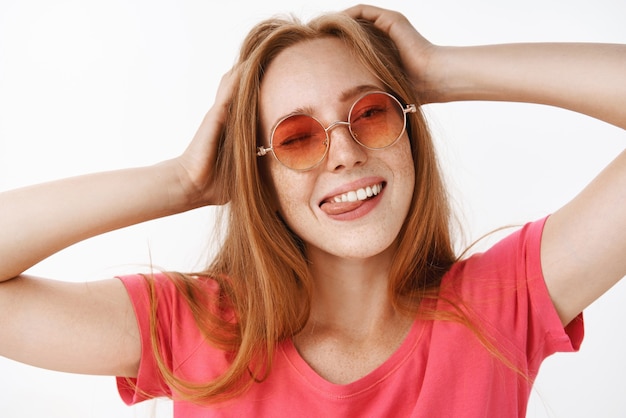 Chica elegante con gafas de sol de moda y camiseta rosa cogidos de la mano en la cabeza guiñando un ojo, sacando la lengua felizmente y sonriendo de alegría y emociones relajadas divirtiéndose, relajándose sobre una pared gris