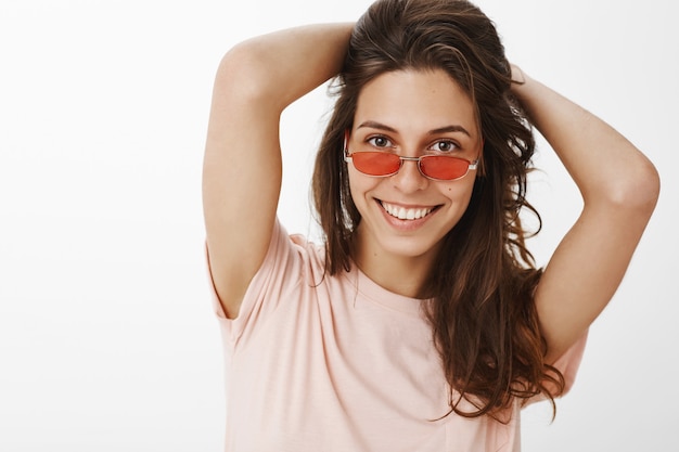 Chica elegante con gafas de sol posando contra la pared blanca