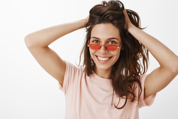 Chica elegante con gafas de sol posando contra la pared blanca