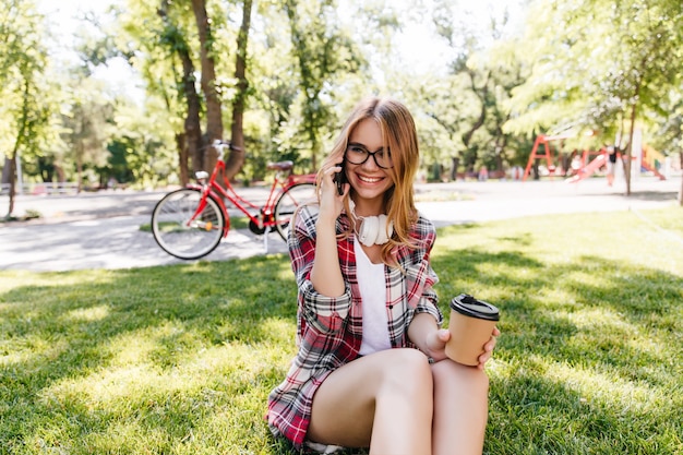 Chica elegante hablando por teléfono mientras está sentado en la hierba. Foto exterior de mujer adorable sonriente con taza de café.