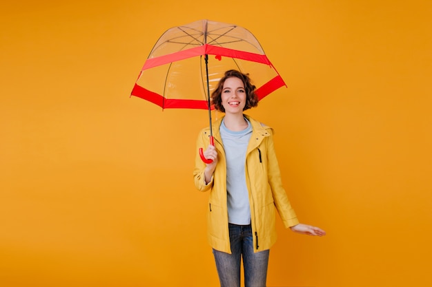 Chica elegante en jeans y gabardina de pie bajo un lindo paraguas. Retrato de interior de mujer joven romántica con peinado rizado con sombrilla en la pared naranja.