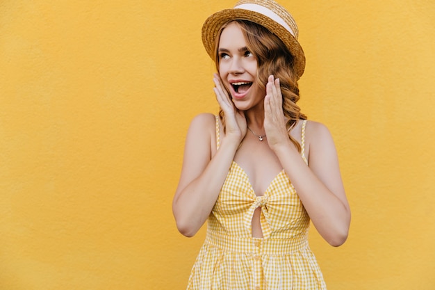 Chica elegante lleva sombrero de paja y collar posando con sonrisa de sorpresa. Mujer rizada asombrada en vestido amarillo de pie.