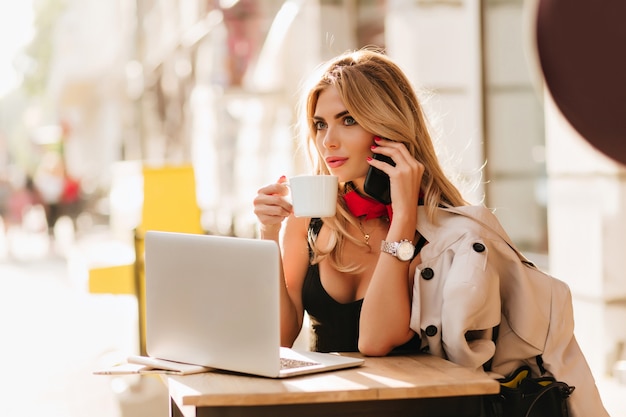 Chica elegante con manicura roja posando con teléfono y taza de café sobre fondo borroso