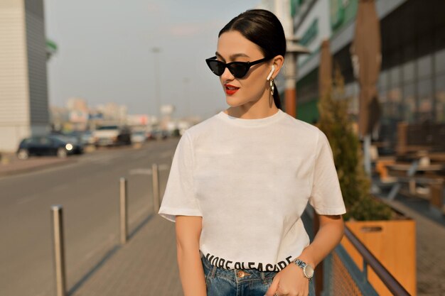 Chica elegante de moda con camiseta blanca y gafas negras con labios rojos está escuchando música a la luz del sol en el fondo de la ciudad