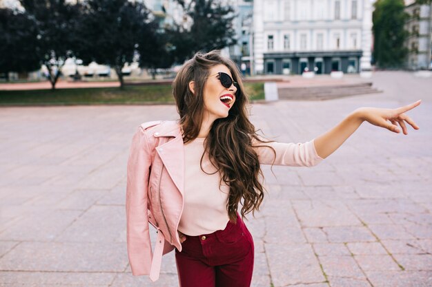 Chica elegante con peinado largo en pantalones vinosos divirtiéndose en la ciudad. Ella tiene chaqueta rosa en el hombro, mostrando a un lado.