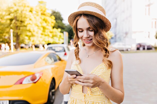 Chica elegante con peinado de moda llamando a alguien mientras está de pie en la calle. Retrato al aire libre de magnífica mujer caucásica con sombrero de paja.