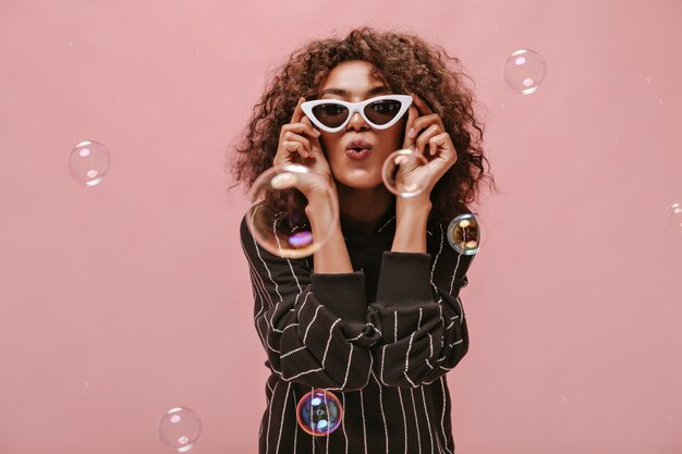 Chica elegante con peinado ondulado corto en traje negro a rayas que sopla beso, sosteniendo gafas de sol blancas y posando con burbuja en la pared rosa.