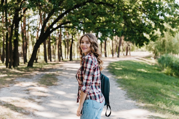 Chica elegante con peinado rizado mirando por encima del hombro y disfrutando de un paseo en el parque. Señora de buen humor en ropa casual posando en la naturaleza.
