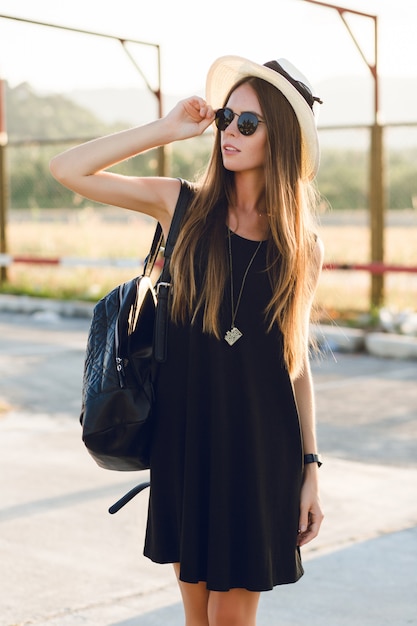 Foto gratuita chica elegante de pie cerca de la carretera con vestido negro corto, sombrero de paja, anteojos negros y mochila negra. ella sonríe bajo los cálidos rayos del sol poniente. ella toca sus gafas de sol con la mano