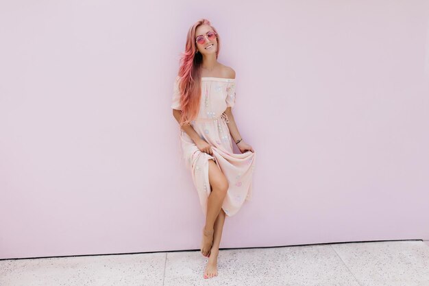 Chica elegante con piel bronceada jugando con su vestido largo sobre fondo blanco Impresionante mujer riéndose con cabello rosado divirtiéndose durante la sesión de fotos en interiores