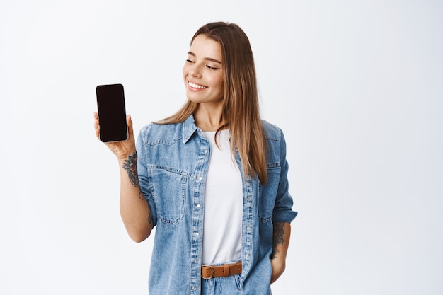 Chica elegante que muestra la pantalla vacía del teléfono móvil y parece complacida, recomendando la aplicación para el teléfono inteligente, demostrando el sitio de compras, pared blanca