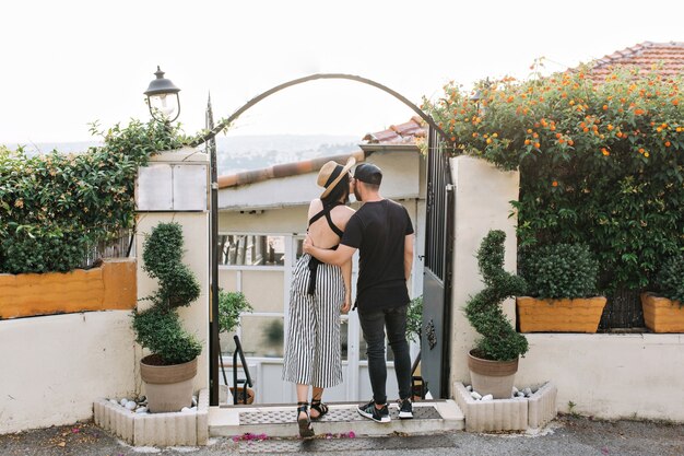 Chica elegante con sombrero besa a su novio de pie frente a puertas negras con plantas exóticas en la mañana