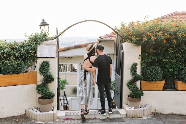 Chica elegante con sombrero besa a su novio de pie frente a puertas negras con plantas exóticas en la mañana