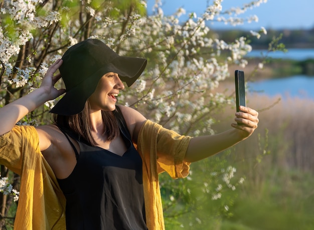 Una chica elegante con un sombrero hace un selfie al atardecer cerca de árboles en flor en el bosque