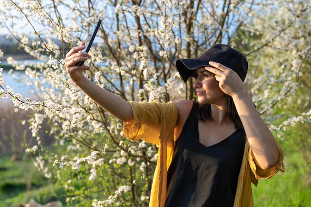 Una chica elegante con un sombrero hace un selfie al atardecer cerca de árboles en flor en el bosque