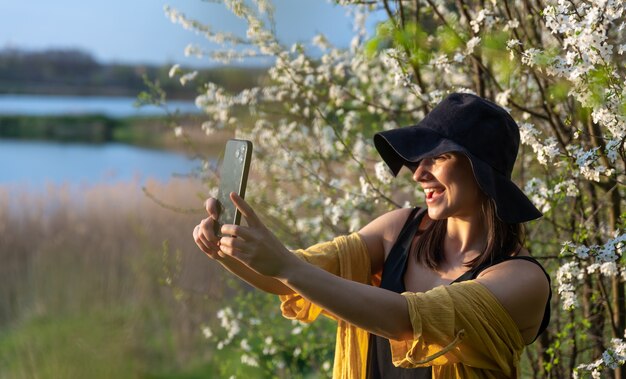 Una chica elegante con un sombrero hace un selfie al atardecer cerca de árboles en flor en el bosque