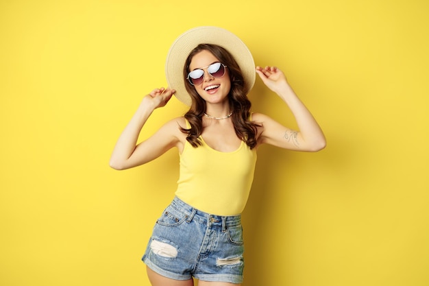 Chica elegante con sombrero de paja y camiseta sin mangas lista para el verano yendo de vacaciones y sonriendo complacida standi...