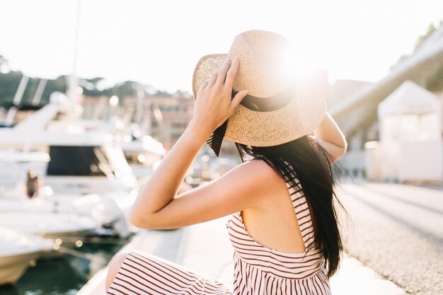 Chica elegante con sombrero con pelo oscuro largo brillante relajante solo, sentado en el puerto por la mañana. Señora joven atractiva en traje de verano escalofriante afuera mirando barcos, disfrutando del tiempo libre.