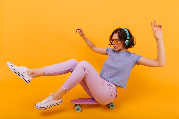 Chica elegante con tatuaje sentado en longboard. Modelo de mujer agradable con el pelo corto y rizado posando en patineta y escuchando música.