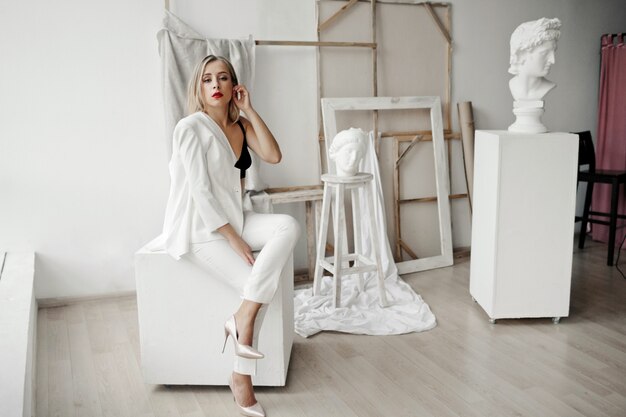 Chica elegante con un traje blanco se sienta en un cubo blanco en una galería