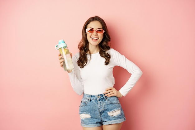 Chica elegante vestida de primavera, con gafas de sol, sosteniendo una botella de agua con limón, bebida saludable, riendo y sonriendo, de pie sobre un fondo rosa.