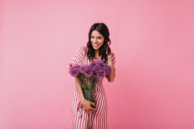 Chica con elegante vestido midi rosa se ve de manera amistosa, posando con un montón de flores para retratos en interiores.