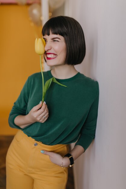 Chica elegante viste pantalones amarillos haciendo muecas mientras posa con tulipán. Impresionante mujer morena con labios rojos sosteniendo flores y riendo.