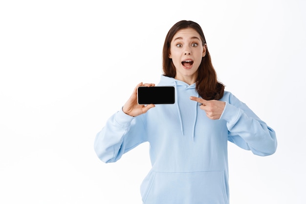 Chica emocionada apuntando y mostrando la pantalla del teléfono inteligente volteada horizontalmente, demuestra una aplicación, de pie contra la pared blanca