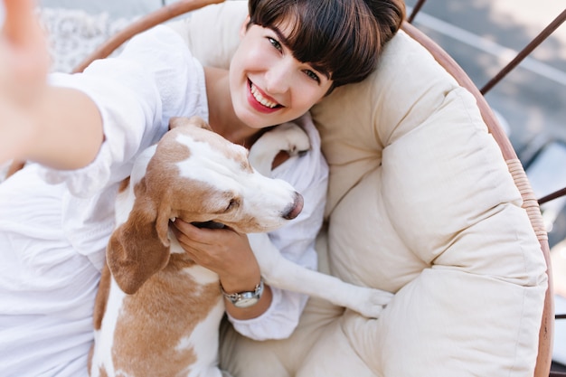 Chica emocionada con cabello castaño corto riendo mientras toma una foto de sí misma con un perro beagle.