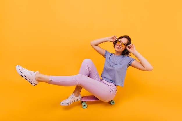 Chica emocionada con gafas de color amarillo sentado en longboard con sonrisa alegre. Señora europea jocund con tatuaje que expresa emociones positivas