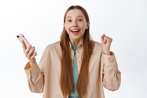 Chica emocionada ganando en línea, saltando de la felicidad con el teléfono inteligente, celebrando la victoria, recibiendo un mensaje con buenas noticias, sonriendo y diciendo que sí, pared blanca