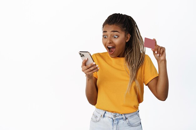 Chica emocionada mirando su teléfono móvil y mostrando la tarjeta de crédito sorprendida por la aplicación bancaria de pie sobre fondo blanco.