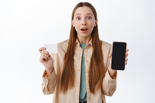 Foto gratuita chica emocionada muestra la pantalla vacía de la tarjeta de crédito y el teléfono móvil, se ve sorprendida, hace un pedido por internet, compra en línea, de pie sobre una pared blanca