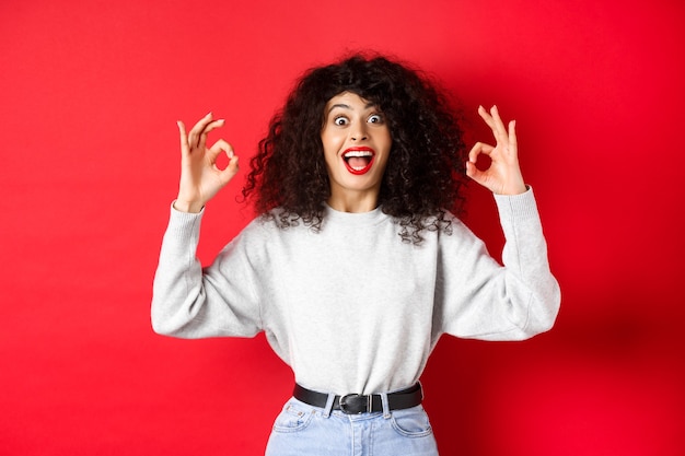 Chica emocionada muestra signos de bien y sonriendo feliz, recomendando un buen artículo, alabando la elección, de pie asombrada sobre fondo rojo