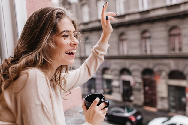 Chica emocionada con peinado rizado agitando la mano a alguien, mirando la ventana