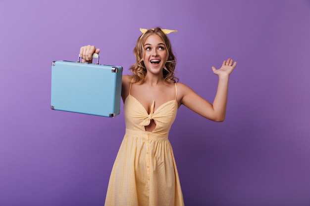 Chica emocionada con piel bronceada sosteniendo su maleta de viaje. Riendo mujer dichosa con maleta expresando emociones positivas.