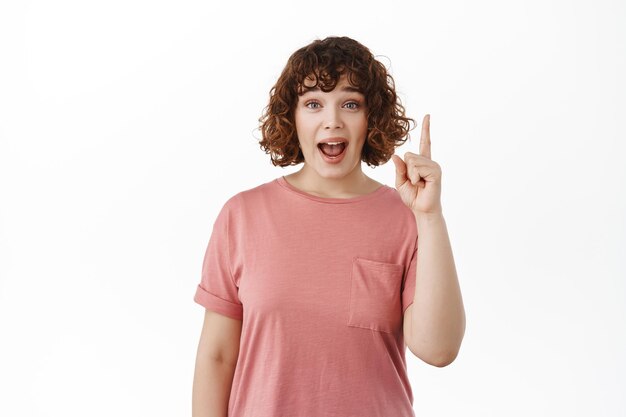 Una chica emocionada y sorprendida grita de asombro, señala con el dedo hacia arriba, dice wow, mira el anuncio en la parte superior, se para en una camiseta contra fondo blanco