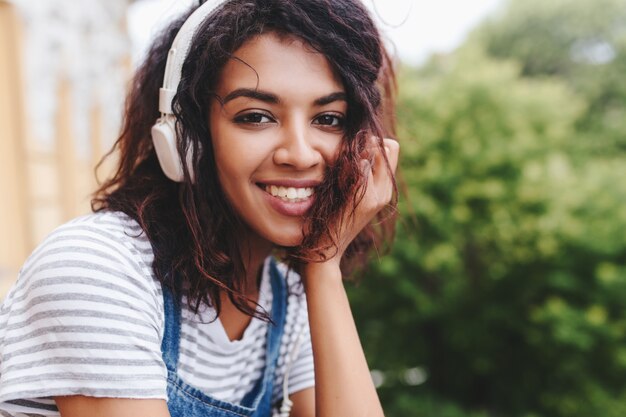 Chica emocionada viste camisa a rayas sentada al aire libre apuntando la cara con la mano y escuchando música con auriculares blancos