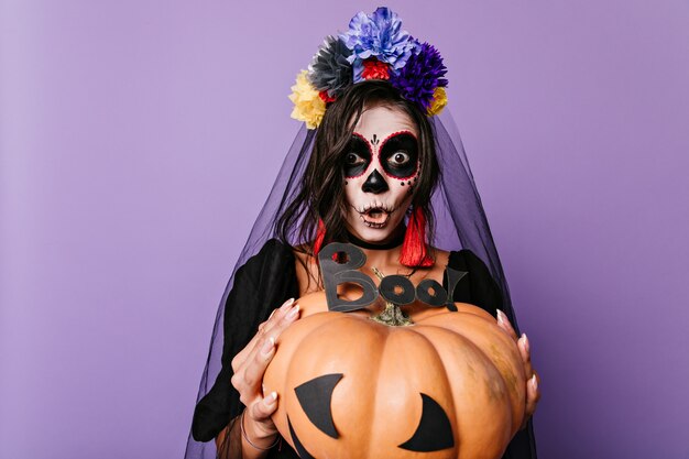 Chica emocional grita "boo" sosteniendo una enorme calabaza decorada frente a ella. Retrato de novia negra pintada en forma de calavera.