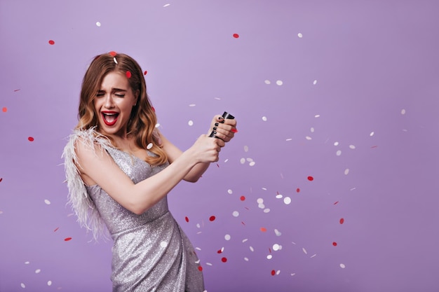 Chica emocional con labios rojos vestida con traje plateado lanzando confeti Mujer emocionada con vestido brillante sonriendo sobre fondo púrpura aislado