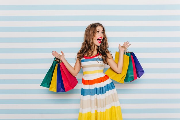 La chica emocional se sorprende por los grandes descuentos durante el viernes negro y compra mucha ropa. Retrato de cuerpo entero de mujer con vestido brillante con bolsas de la compra.