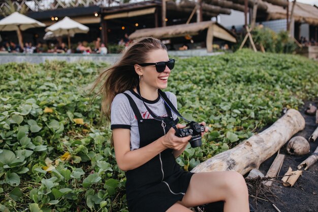 Chica emocional en traje vintage sentado en el césped con cámara. Riendo fotógrafa con gafas de sol negras divirtiéndose en el parque en verano.